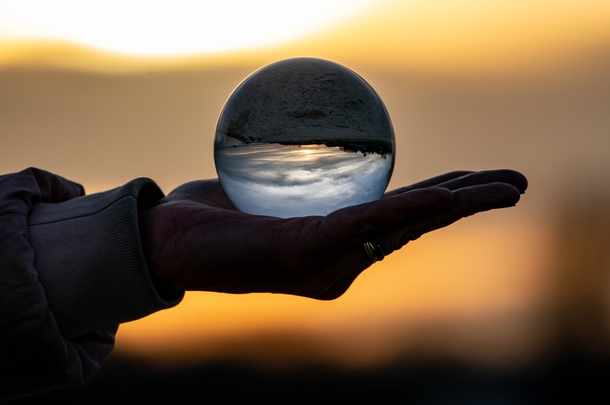 ARCHIV - 20.02.2021, Hamburg, Surendorf: Eine Frau hält am Strand eine Glaskugel in der Hand, in der sich die Umgebung und der Sonnenuntergang spiegeln (gestellte Szene). (zu dpa-Korr &quot;Überall St ...