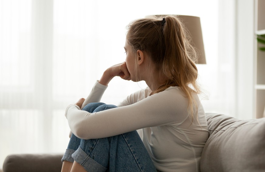 Side view young woman looking away at window sitting on couch at home. Frustrated confused female feels unhappy problem in personal life quarrel break up with boyfriend or unexpected pregnancy concept