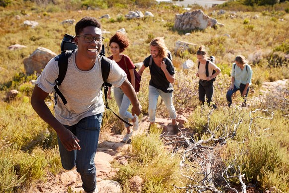 Millennial African American man leading friends hiking single file uphill on a path in countryside