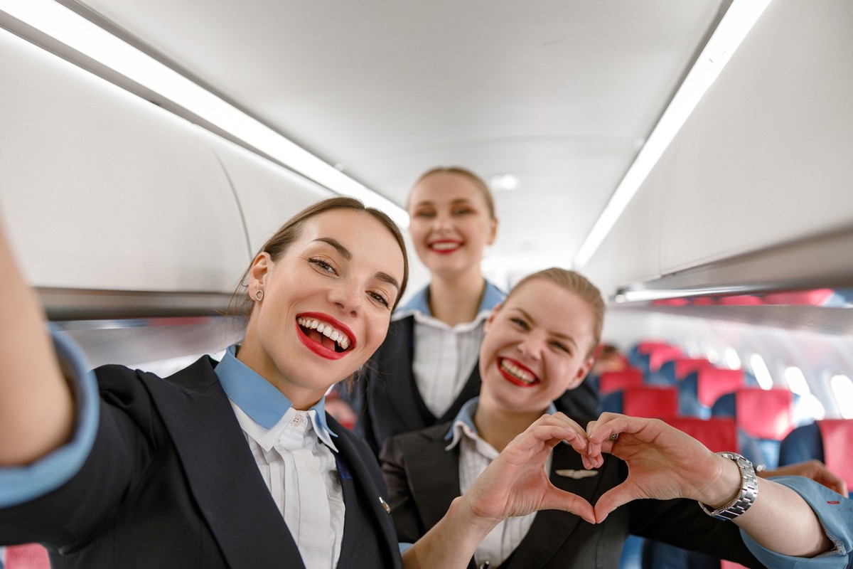 Joyful woman stewardess doing heart symbol with hands and smiling while standing with colleagues in aircraft cabin