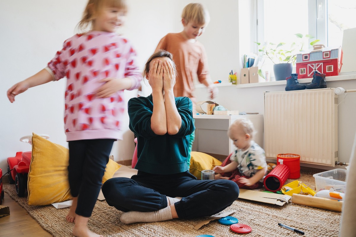 Stressed out mother sitting on floor in middle of toys while children naughty running around her at room. Woman alone burnout with kids. Family home with chaos, mess. Motion blur for speed, real life.