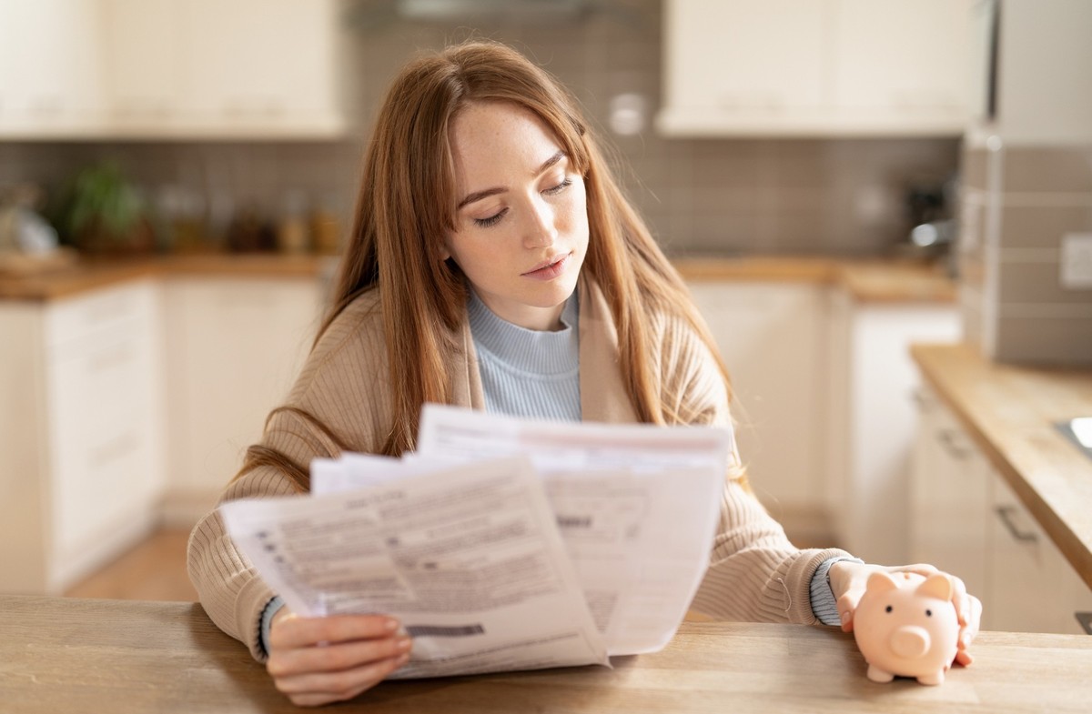 A woman sits at a kitchen table examining bills and financial papers while holding a piggy bank, focusing intently on her finances in a cozy space.