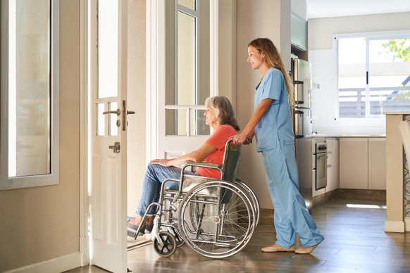Pflegekraft oder Krankenschwester schiebt eine gelähmte Seniorin im Rollstuhl im Seniorenheim || Modellfreigabe vorhanden