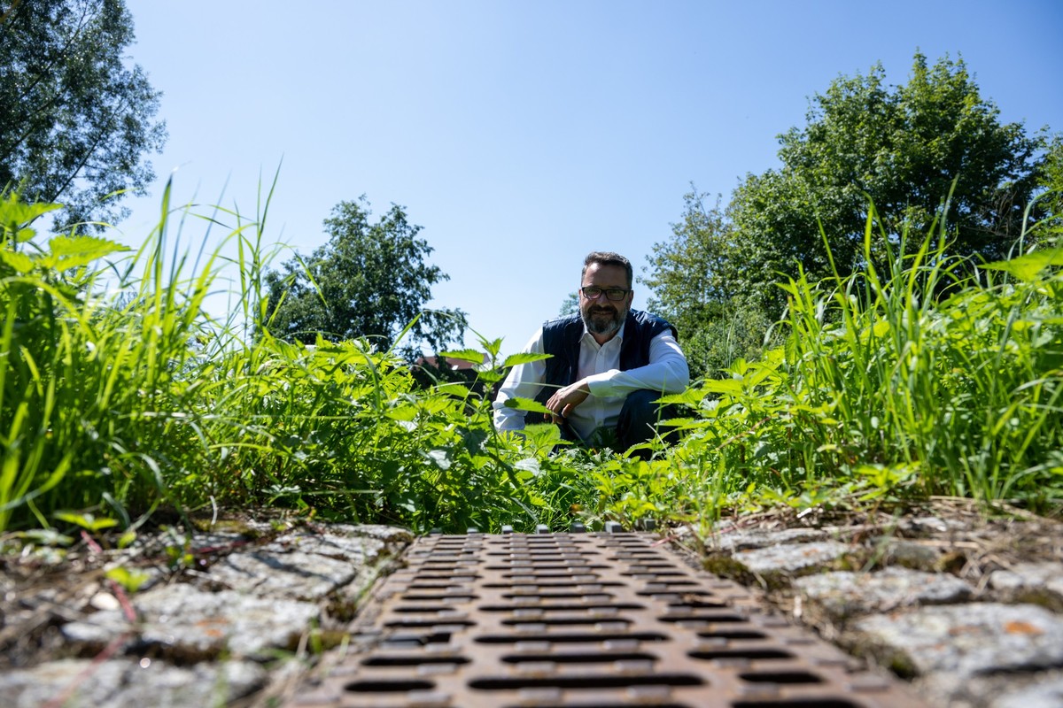PRODUKTION - 11.08.2023, Bayern, Neu-Ulm: Jochen Meissner, Leiter der Abteilung Stadtentwässerung und Wasserbau der Stadt Neu-Ulm, kniet hinter der Regenrinne einer Straße, die in einem Regenrücklaufb ...