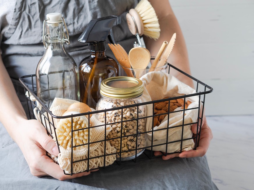 Women hold black metal basket with eco-friendly kitchen set. Food, brushes, wood appliances, bags, bottle and jar.