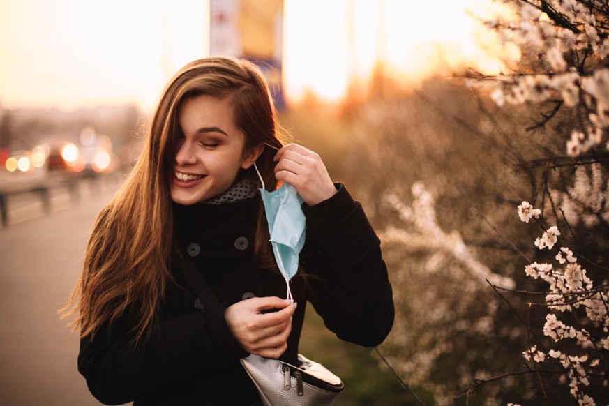 Happy cheerful young woman removing face medical mask while standing on street in city during sunset in spring